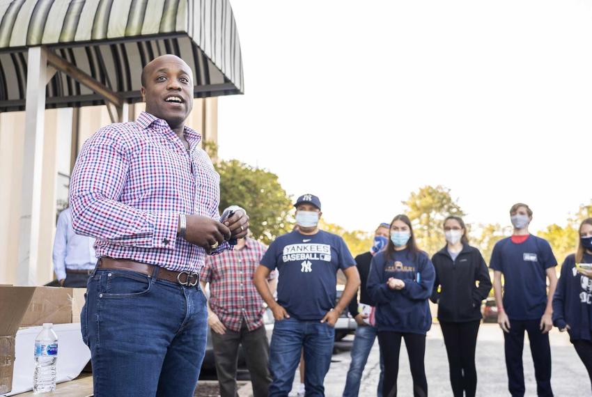 A Black man in a plaid shirt and jeans stands outside in front of a row of casually dressed people wearing face masks.