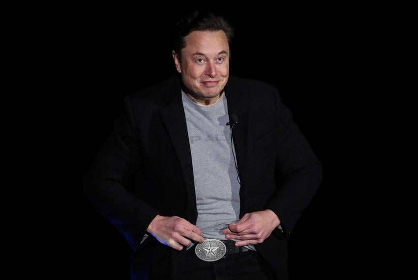 Musk, wearing a black blazer and a gray SpaceX T-shirt, lifts the hem of his shirt to show a large silver belt buckle featuring a five-pointed star with Tesla’s stylized “T” logo in the middle.